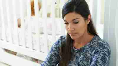 An overwhelmed mom sits by her child's crib and looks sad.