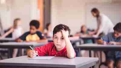 Portrait of bored schoolboy in red shirt sitting at a desk and looking frustrated, zoned out, unfocused, inattentive.