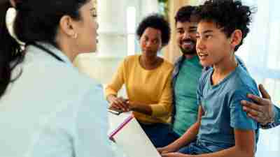 An female Asian pediatrician gives an in-person ADHD diagnosis to a young black boy with his mom and dad smiling in the background, which would not be seen during a telehealth ADHD evaluation.