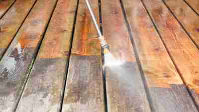 A pressure washer sprayer is cleaning a weathered treated wood deck. The background wood has been cleaned, the lower portion is dirty and weathered, showing the contrast. Focus is on the wood, the nozzle is slightly soft.