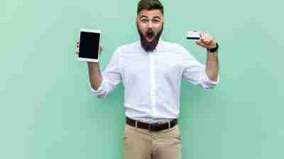 No way! Online shopping. Businessman holding hands credit card and tablet and looking at with shocked face. Indoor, studio shot. Isolated on light green background