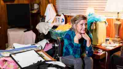 A senior woman in her 60s at home, sitting in a messy, cluttered room, looking away with a serious expression.