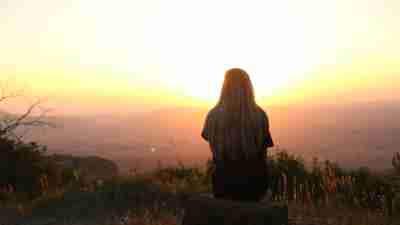 A pensive woman sitting on a rock on top of a mountain and looking at the sunset, reflecting on imposter syndrome, feelings of inadequacy, RSD, and ADHD